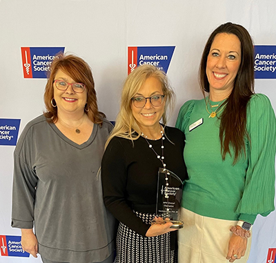 three women standing in front of a American Cancer Society backdrop, middle lady is holding her award